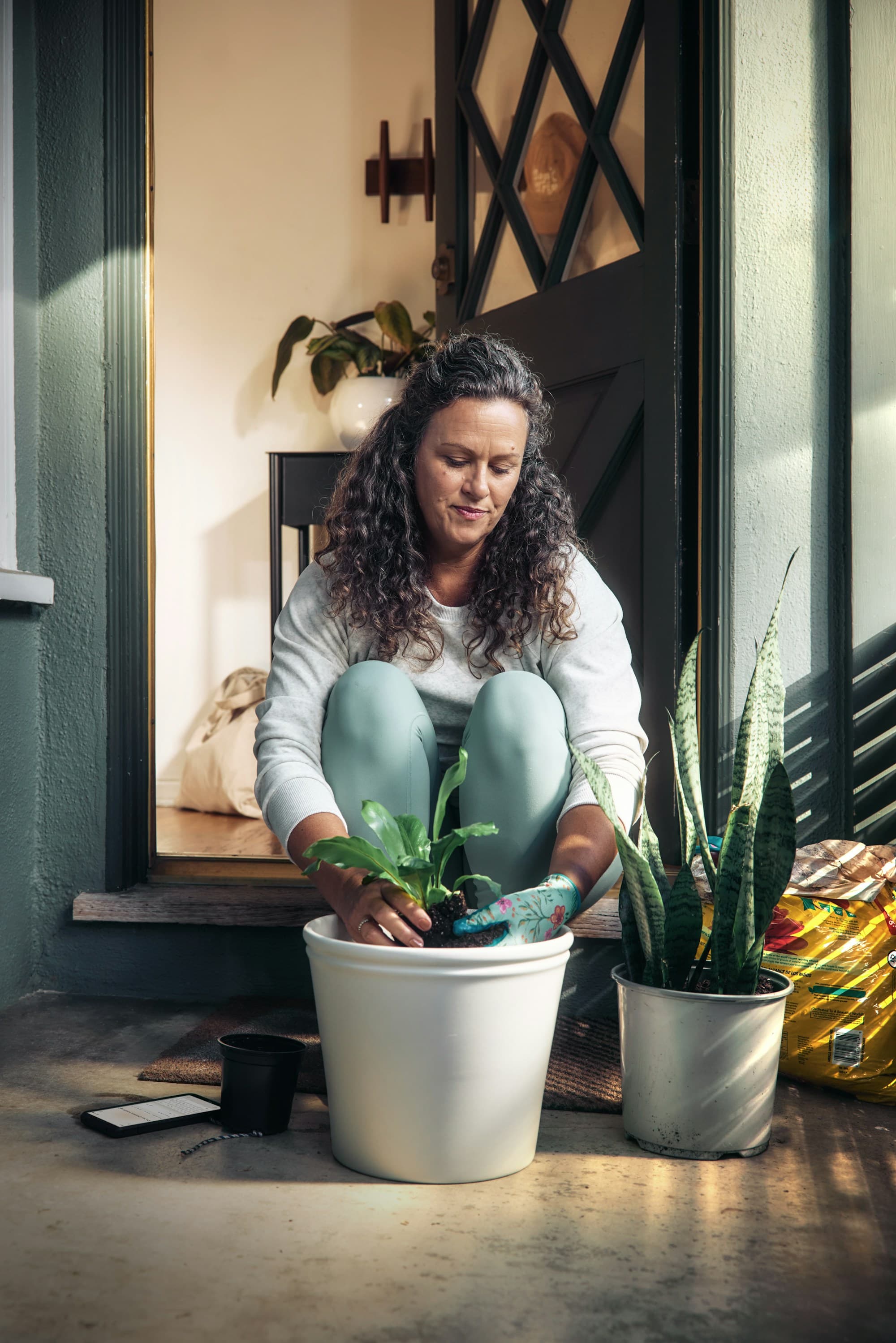 A woman repotting a houseplant on her porch step, a generic-brand potting mix bag beside her